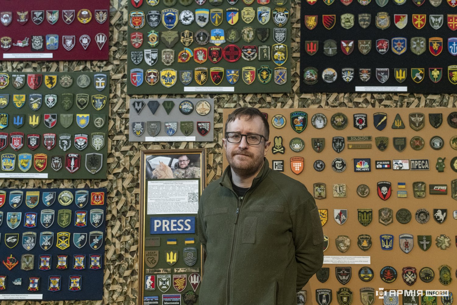 Стів Андре на тлі зібраної ним колекції нарукавних знаків під час відкриття виставки у Національний військово-історичний музей України. За його спиною розміщені стенди з десятками шевронів і патчів різних підрозділів Сил оборони України та суміжних структур.