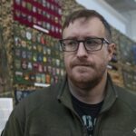 Стів Андре на тлі зібраної ним колекції нарукавних знаків під час відкриття виставки у Національний військово-історичний музей України. За його спиною розміщені стенди з десятками шевронів і патчів різних підрозділів Сил оборони України та суміжних структур. Колекція впорядкована за тематикою й оформлена у вигляді музейної експозиції, що дозволяє розглянути кожен знак окремо. Образ Стіва Андре на фоні експонатів підкреслює особисту причетність колекціонера до створення цієї унікальної виставки.