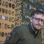 Стів Андре на тлі зібраної ним колекції нарукавних знаків під час відкриття виставки у Національний військово-історичний музей України. За його спиною розміщені стенди з десятками шевронів і патчів різних підрозділів Сил оборони України та суміжних структур. Колекція впорядкована за тематикою й оформлена у вигляді музейної експозиції, що дозволяє розглянути кожен знак окремо. Образ Стіва Андре на фоні експонатів підкреслює особисту причетність колекціонера до створення цієї унікальної виставки.