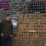 Стів Андре на тлі зібраної ним колекції нарукавних знаків під час відкриття виставки у Національному військово-історичному музеї України. За його спиною розміщені стенди з десятками шевронів і патчів різних підрозділів Сил оборони України та суміжних структур. Колекція впорядкована за тематикою й оформлена у вигляді музейної експозиції, що дозволяє розглянути кожен знак окремо. Образ Стіва Андре на фоні експонатів підкреслює особисту причетність колекціонера до створення цієї унікальної виставки.