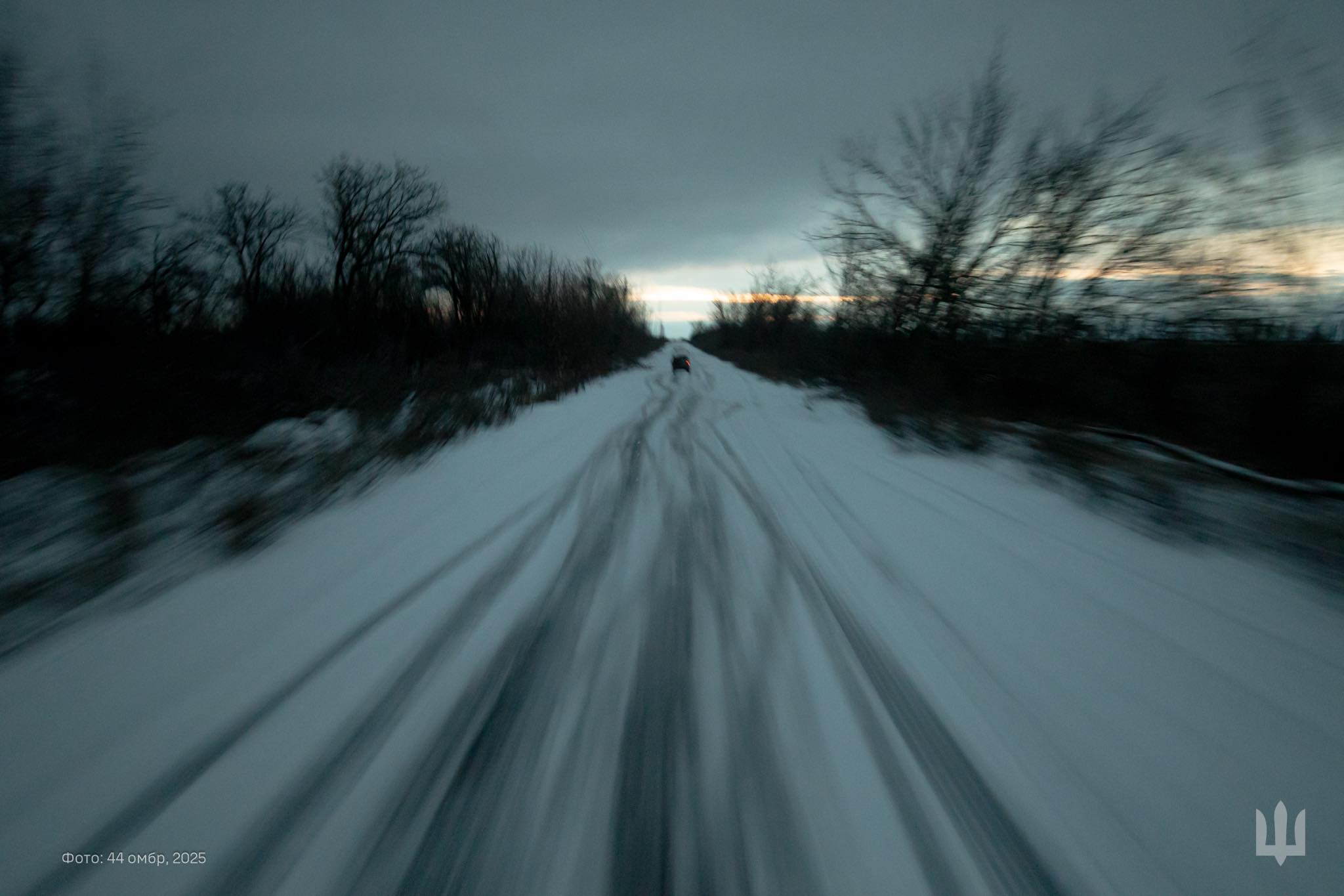 На фото — засніжена польова дорога, що тягнеться вперед між темними деревами та кущами. Знімок зроблено в русі: через розмиття видно колії на снігу й силует автомобіля вдалині. Над горизонтом нависли важкі хмари, а вузька смуга світла на заході додає кадру холодної напруги.