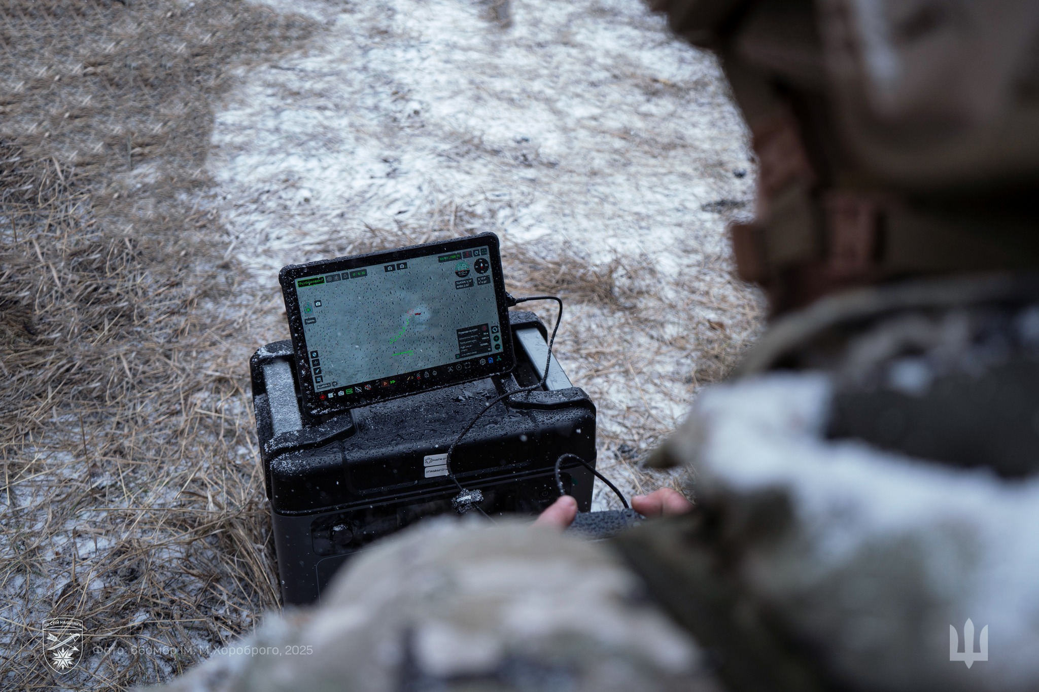 На фото військовослужбовець працює з обладнанням керування дроном у польових умовах. На екрані планшета відображається карта та інтерфейс моніторингу польоту. Навколо видно зимовий ґрунт зі снігом і сухою травою, а техніка вкрита краплями вологи.