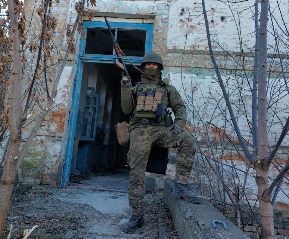 Український захисник у повному спорядженні стоїть біля пошкодженої будівлі зі зброєю в руках.