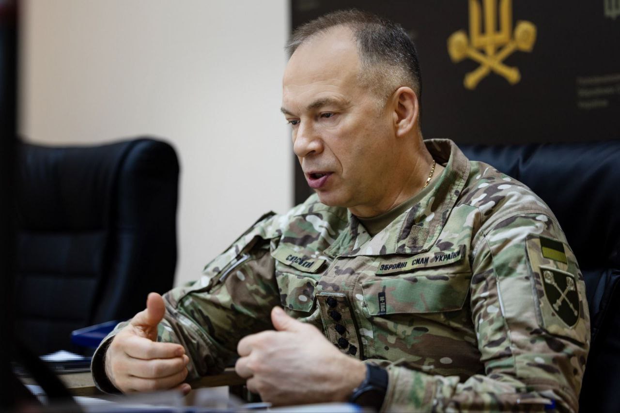 Портрет Головнокомандувача ЗСУ Олександра Сирського під час робочої наради чи інтерв'ю. Генерал одягнений у військову форму (мультикам) із нашивками «Збройні Сили України» та іменним прізвищем. Він сидить за столом, жестикулюючи руками під час пояснення важливих питань. Погляд зосереджений, вираз обличчя серйозний. На задньому плані видно частину чорного банера з золотистою військовою символікою.