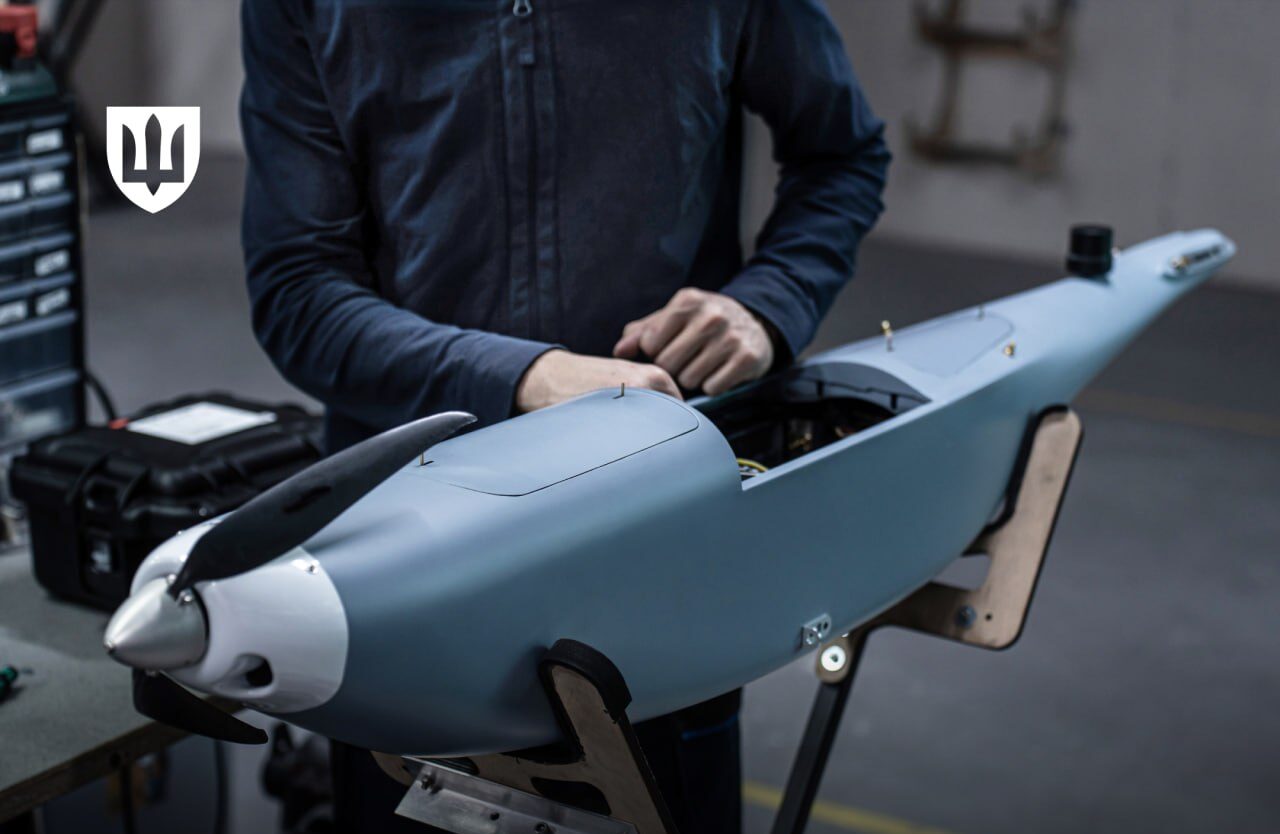 Фотографія з майстерні або лабораторії, де людина в синьому одязі працює над складанням великого безпілотника сіро-блакитного кольору. БпЛА має обтічну форму та великий пропелер у носовій частині. У верхньому лівому куті — емблема Міністерства оборони України. У нижній частині зображення на чорному фоні розміщено великий білий та золотистий напис: «1 млрд грн на пільгове кредитування та лізинг для зброярів