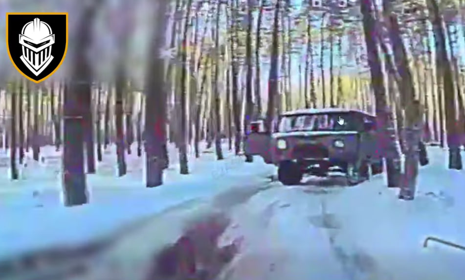 Кадр із камери дрона (низька роздільна здатність, цифрові перешкоди), що зафіксував темний мікроавтобус (типу УАЗ «Буханка») серед засніженого соснового лісу. У верхньому лівому куті — емблема Залізної бригади: білий лицарський шолом на чорному щиті з помаранчевим контуром. Зображення передає момент спостереження за ворожою ціллю в зимових умовах.