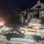 На вулиці зафіксовано пошкоджені транспортні засоби та розкидані уламки. Один автомобіль стоїть біля будівлі з пошкодженими конструкціями. Зйомка виконана вночі.