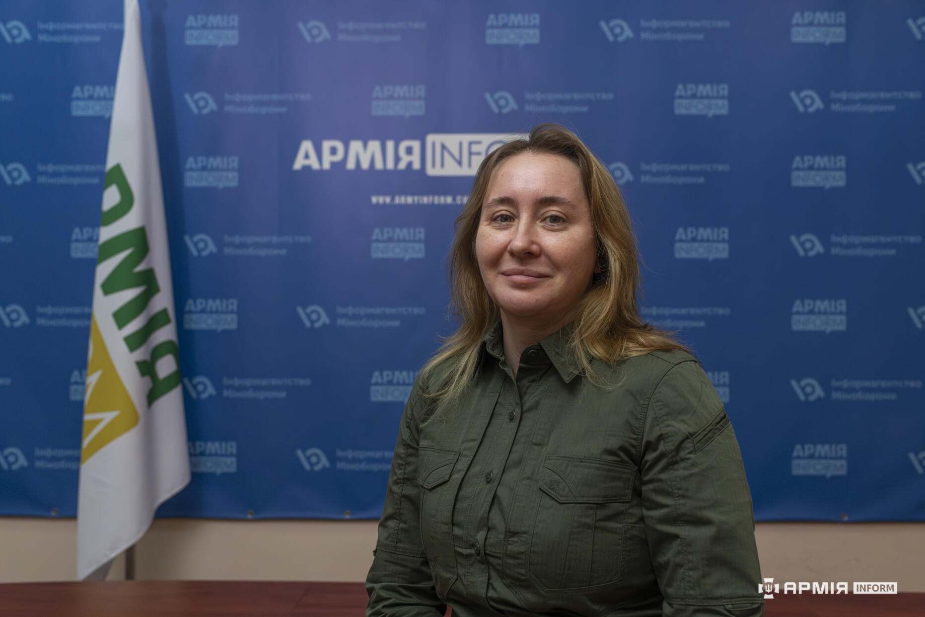 Жінка в оливковій сорочці на фоні брендованої стіни інформаційного агентства «АрміяInform».