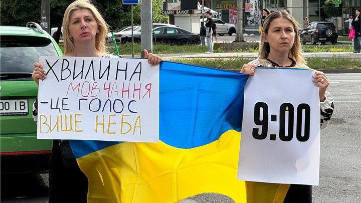 Дві жінки тримають український прапор і плакати із написами про хвилину мовчання та час 9:00. Вони стоять біля дороги, наголошуючи на важливості щоденного вшанування пам’яті.