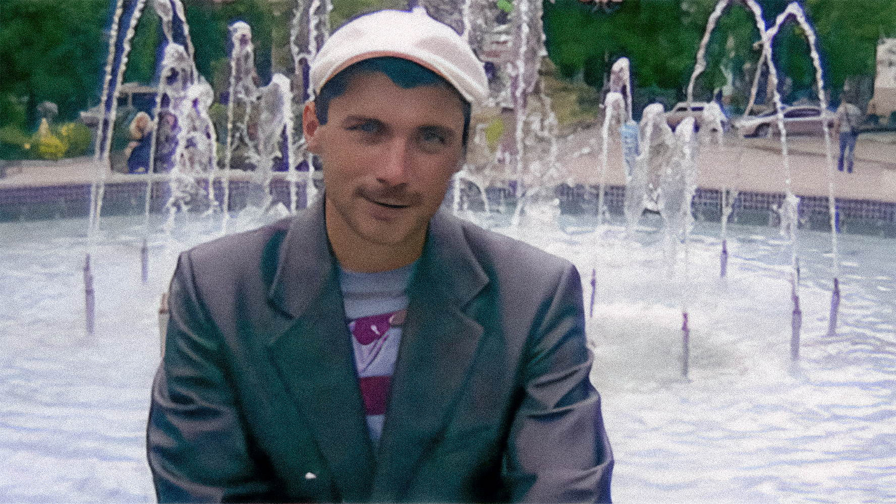 Чоловік у світлій кепці та піджаку сидить біля міського фонтану, за спиною видно струмені води й перехожих. Він дивиться в камеру з легким усміхом, а кадр має трохи зернисту, аматорську якість.