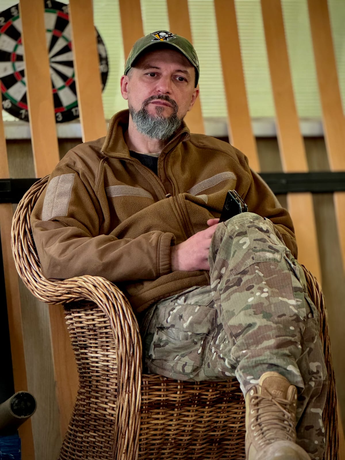 Чоловік у камуфляжних штанах і флісі сидить у плетеному кріслі в приміщенні, тримаючи в руках телефон. Позаду видно мішень для дартсу та вертикальні дерев’яні панелі, що надає кадру неформального інтерв’ю або відпочинку