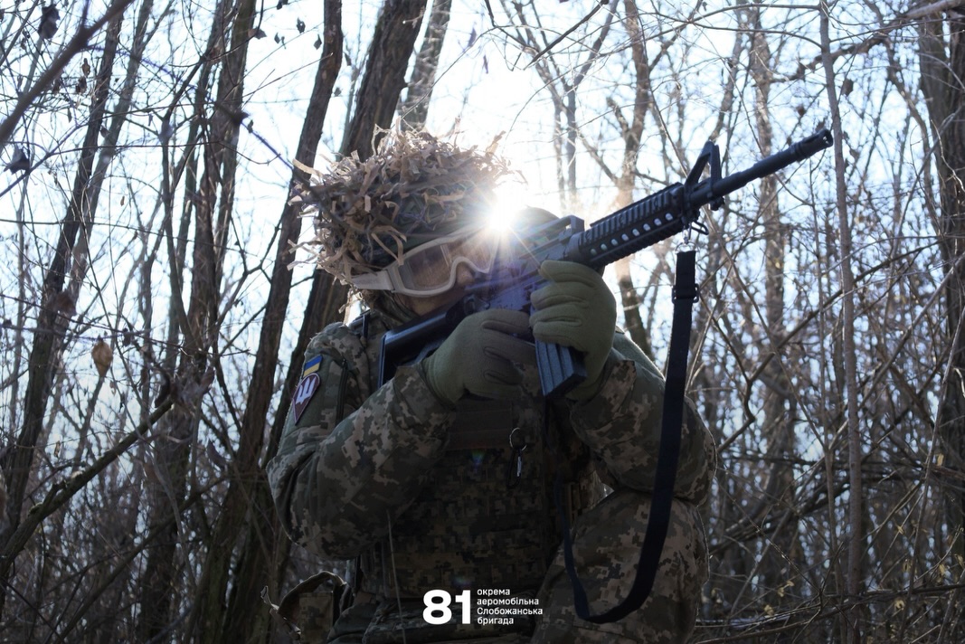 Боєць у камуфляжі та захисних окулярах піднімає автомат і уважно прицілюється серед дерев. Сонце світить позаду нього, створюючи яскравий відблиск і силуетну напруженість сцени.