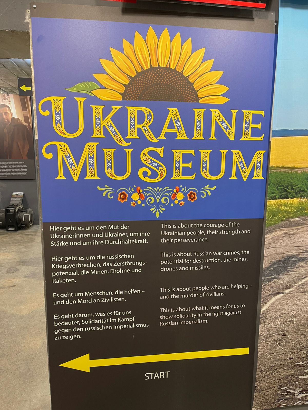Великий стенд із написом “Ukraine Museum” прикрашений соняшником і декоративними орнаментами у синьо-жовтих кольорах. Унизу подано пояснювальний текст про мужність українців, воєнні злочини та міжнародну солідарність.