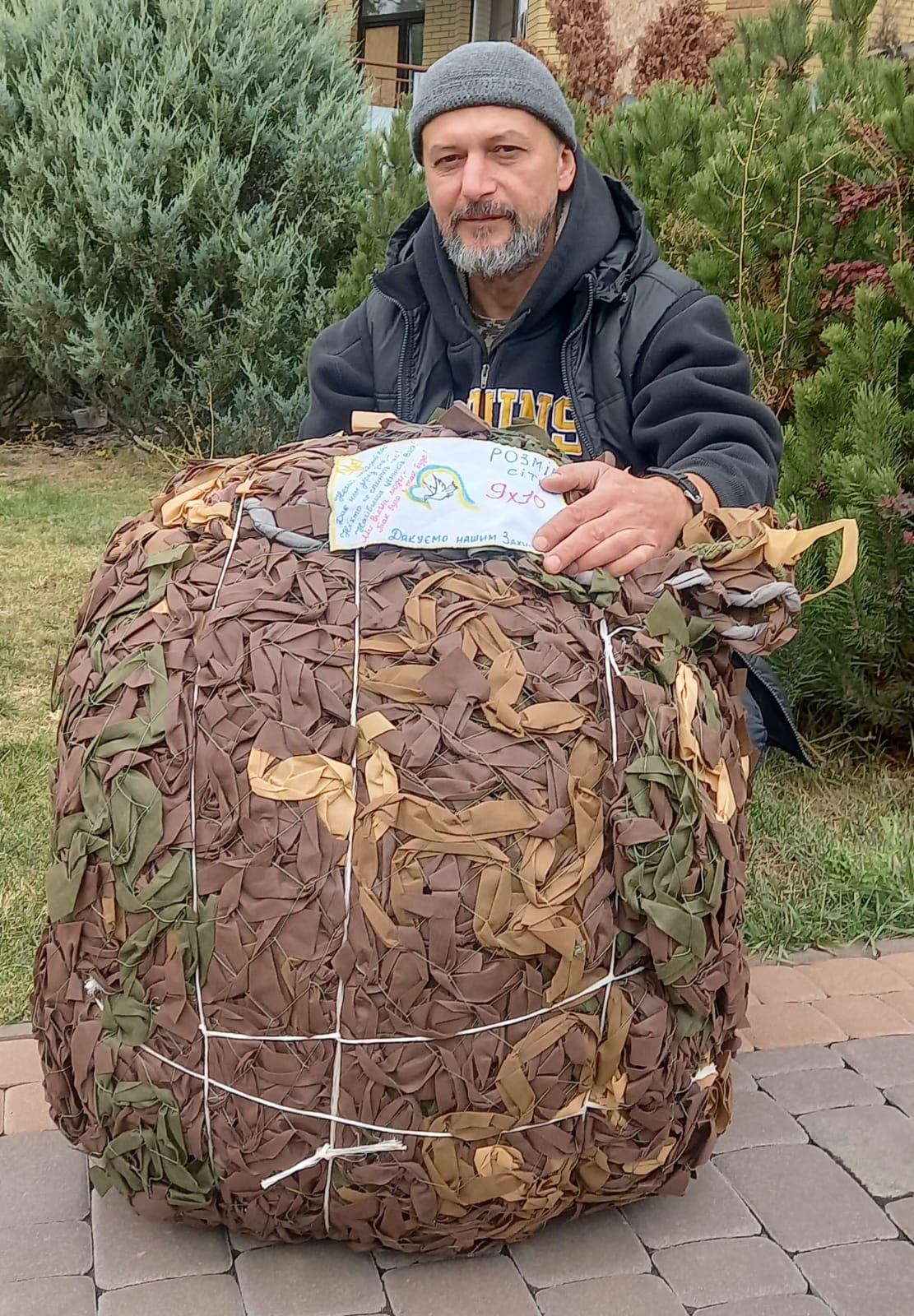 Чоловік у шапці та темному одязі стоїть на подвір’ї й тримає великий пакунок із маскувальною сіткою, перев’язаний мотузками. На пакунку прикріплена дитяча листівка з подякою військовим, що підкреслює волонтерський або гуманітарний контекст.