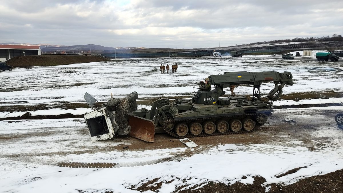 На засніженому полі інженерна броньована машина працює біля перекинутого вантажного автомобіля. Довкола розташована військова техніка та кілька військових у віддаленні.