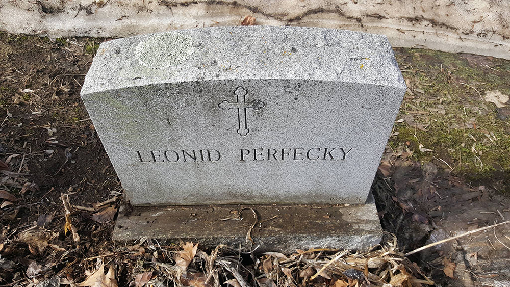 Кам’яний надгробок із викарбуваним написом «LEONID PERIFECKY» і хрестом над ім’ям. Пам’ятник стоїть на землі серед листя та трави.