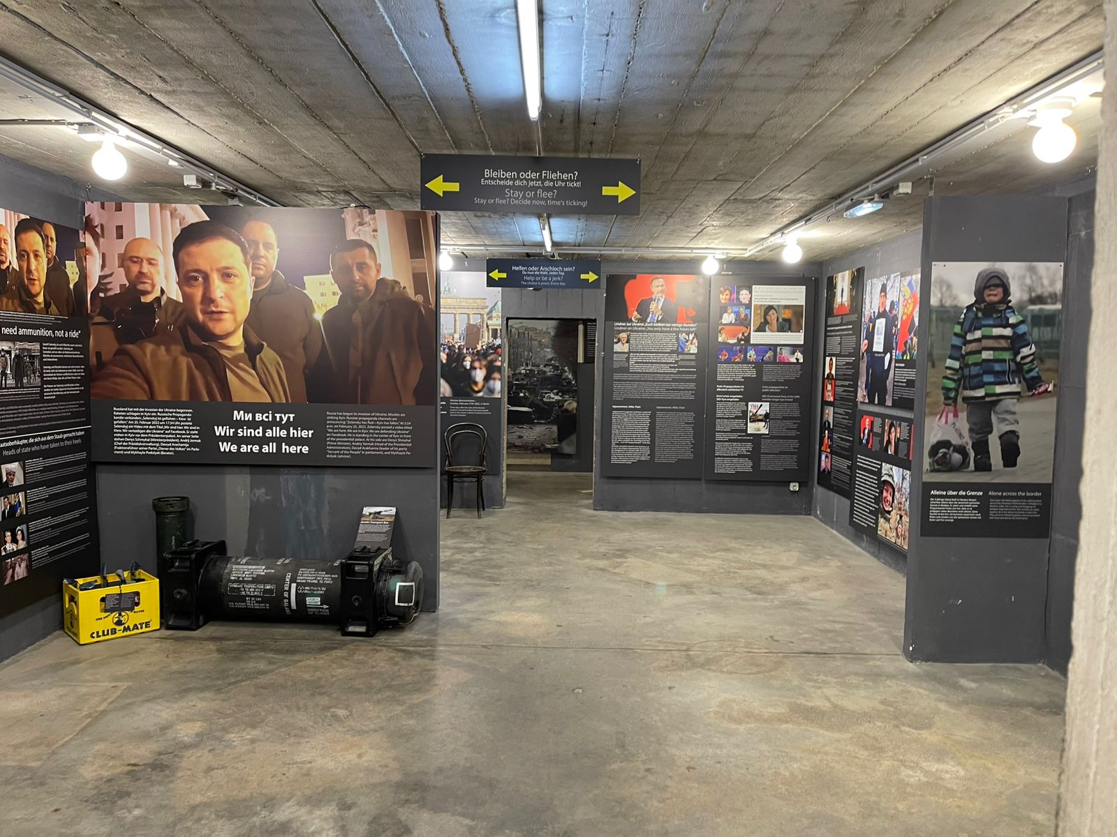 Широкий коридор підземної експозиції заповнений інформаційними панелями з фото, текстами та відеоекранами про війну й гуманітарні історії. На одній зі стін видно великий портрет Володимира Зеленського з підписом про єдність.