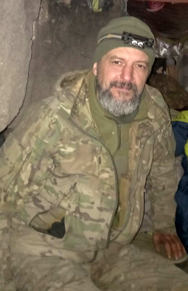 Чоловік у камуфляжі та теплій шапці сидить у тісному підземному або польовому укритті й дивиться в камеру з легкою усмішкою. Тьмяне освітлення й грубі стіни навколо створюють враження фронтових умов.