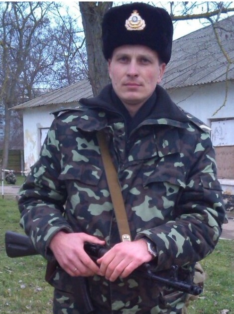 Військовий у камуфляжі та зимовій шапці стоїть надворі біля будівлі, тримаючи в руках автомат. На задньому плані видно подвір’я з деревами та господарськими спорудами, що створює враження службового або польового знімка.