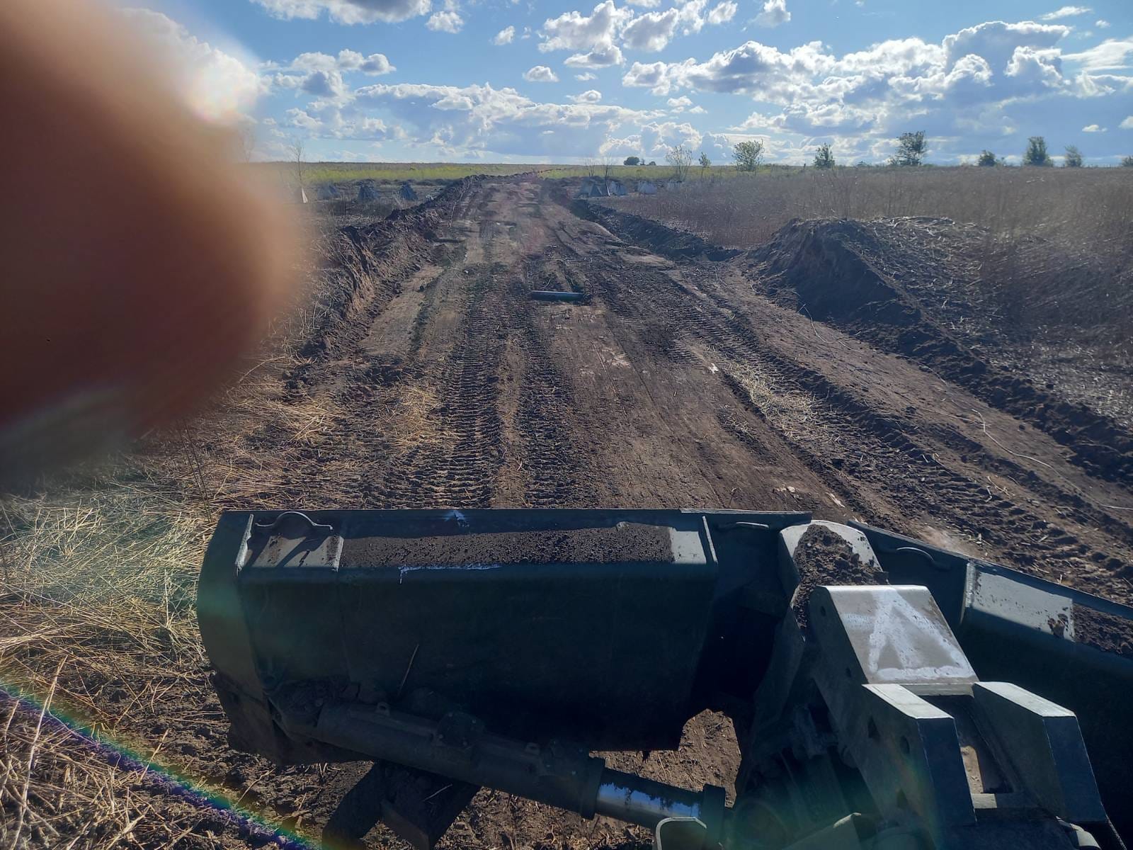 Вид із машини на польову дорогу з виритими валами по боках. Попереду видно сліди техніки, відкритий простір і небо з хмарами, а в кадр частково потрапив палець фотографа.