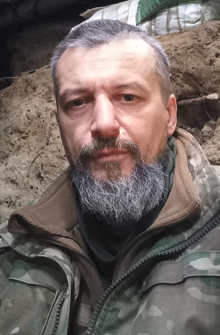 Крупний селфі-портрет показує чоловіка у військовій куртці на фоні земляних укріплень і мішків із ґрунтом. Його серйозний вираз обличчя та польове спорядження вказують на перебування в оборонній позиції.