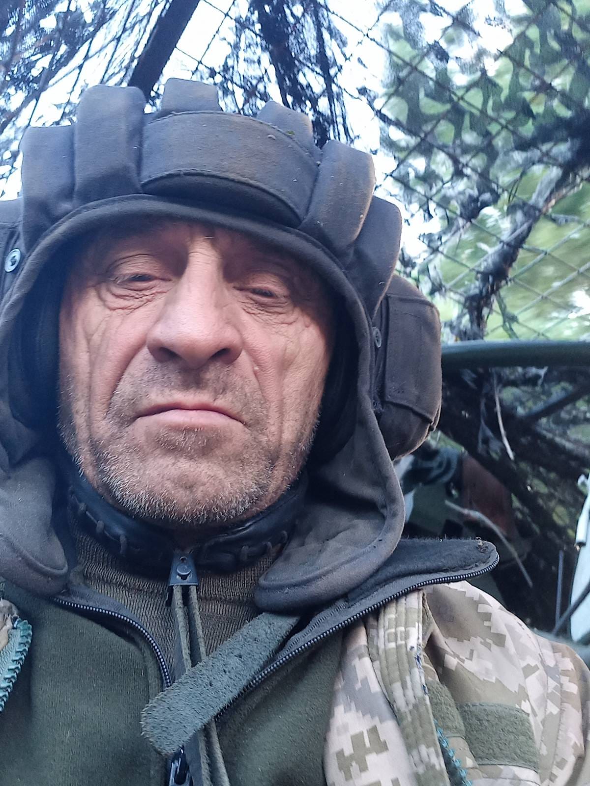 Селфі військового у захисному шоломі та теплому одязі зроблене поруч із технікою та маскувальною сіткою. Обличчя крупним планом, на задньому плані видно елементи позиції.