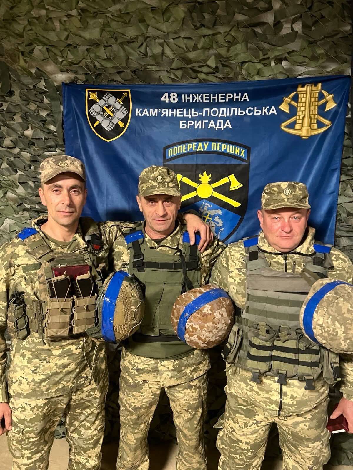 Троє військових у формі стоять разом перед прапором 48-ї інженерної Кам’янець-Подільської бригади. Вони тримають шоломи та позують для спільного знімка в приміщенні з маскувальним тлом.