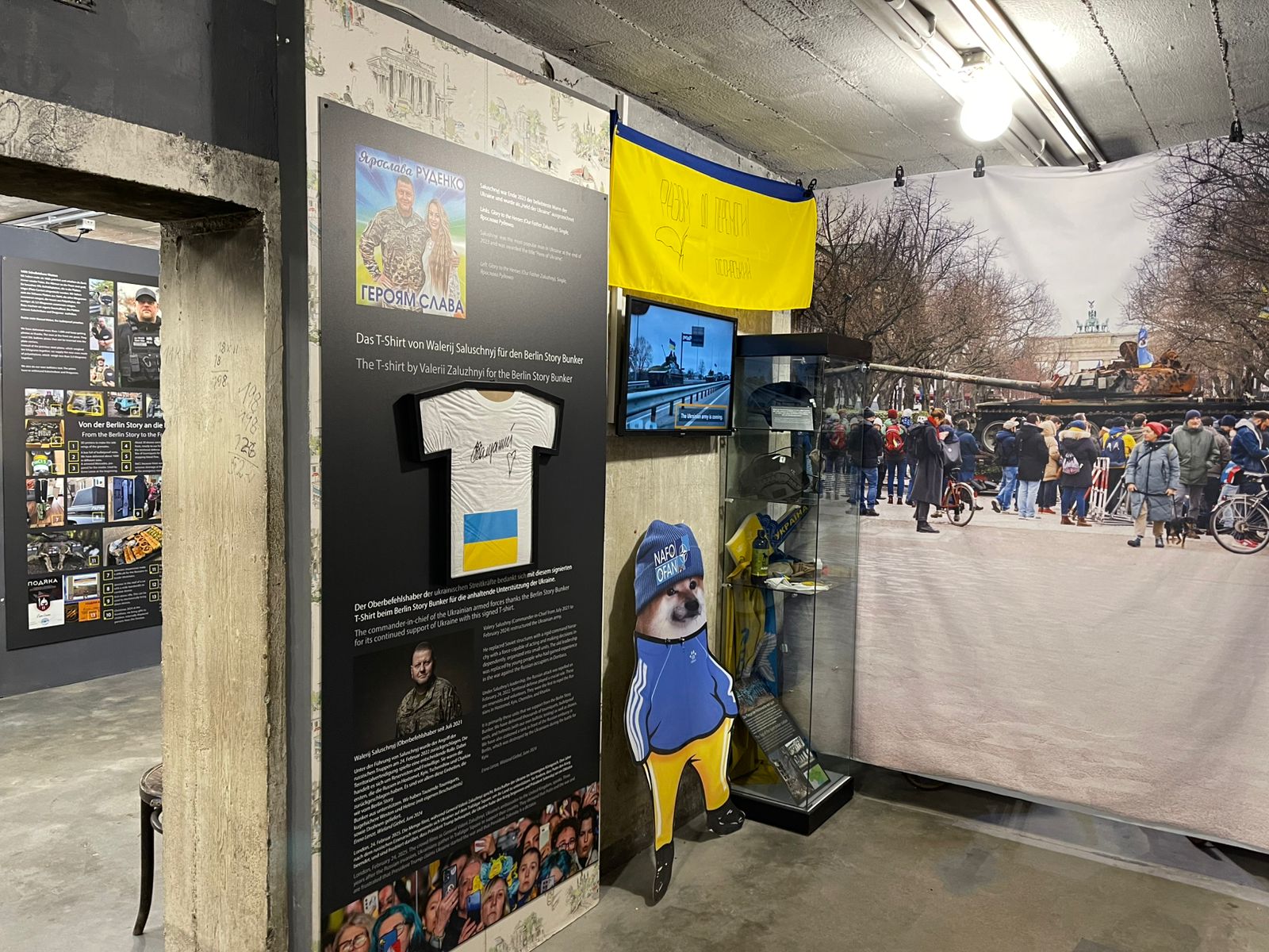 Кут експозиції містить український прапор, підписану футболку, інформаційні стенди й вітрину з пам’ятними речами. Поряд розміщено велике фото з бронетехнікою та натовпом людей, що створює контекст подій у Європі.