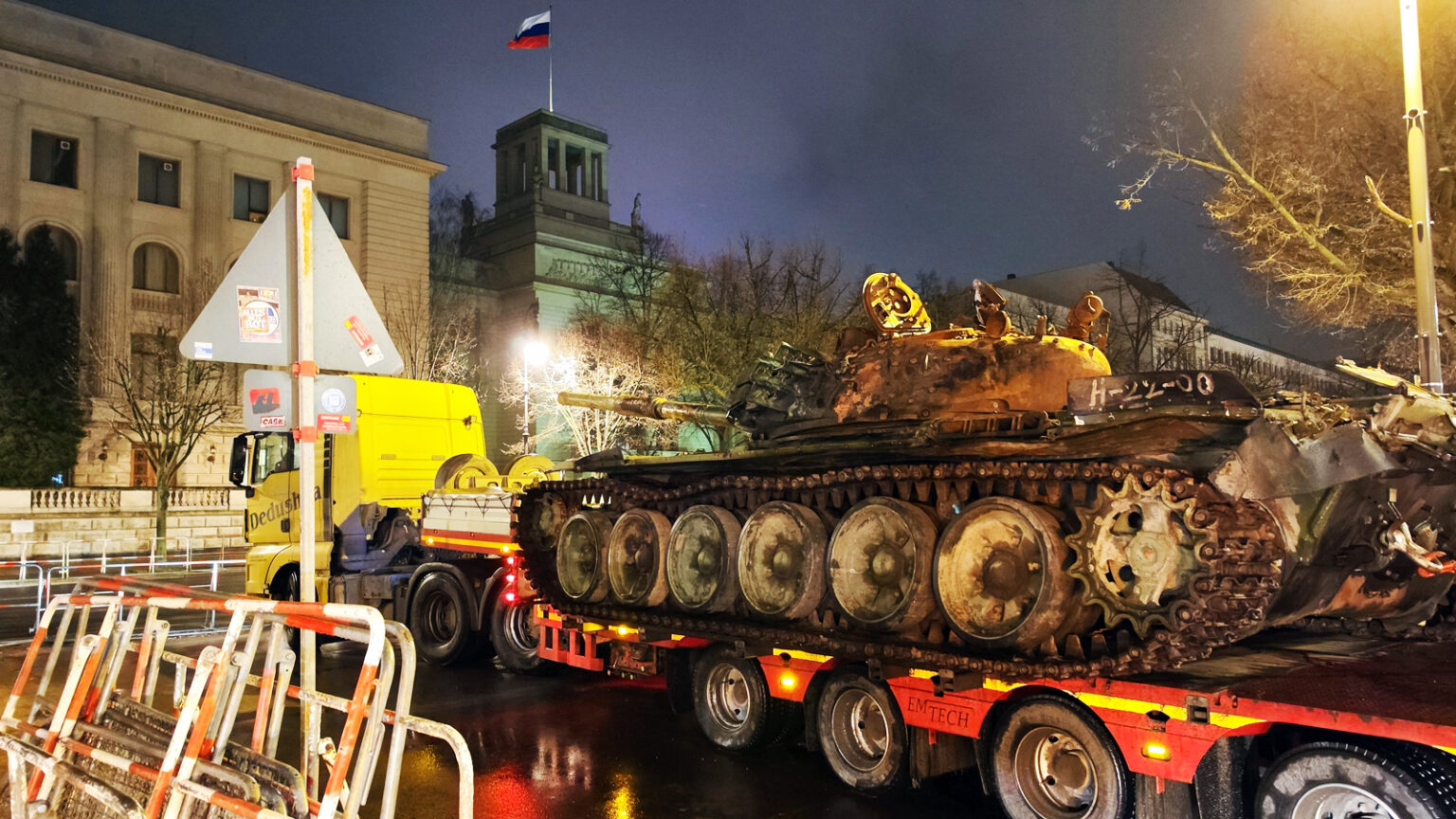 Уночі вулицею перевозять на тралі обгорілий танк, поруч видно міські будівлі та прапор росії на щоглі. Мокрий асфальт відбиває світло ліхтарів і фар, підкреслюючи іржавий стан бойової машини.