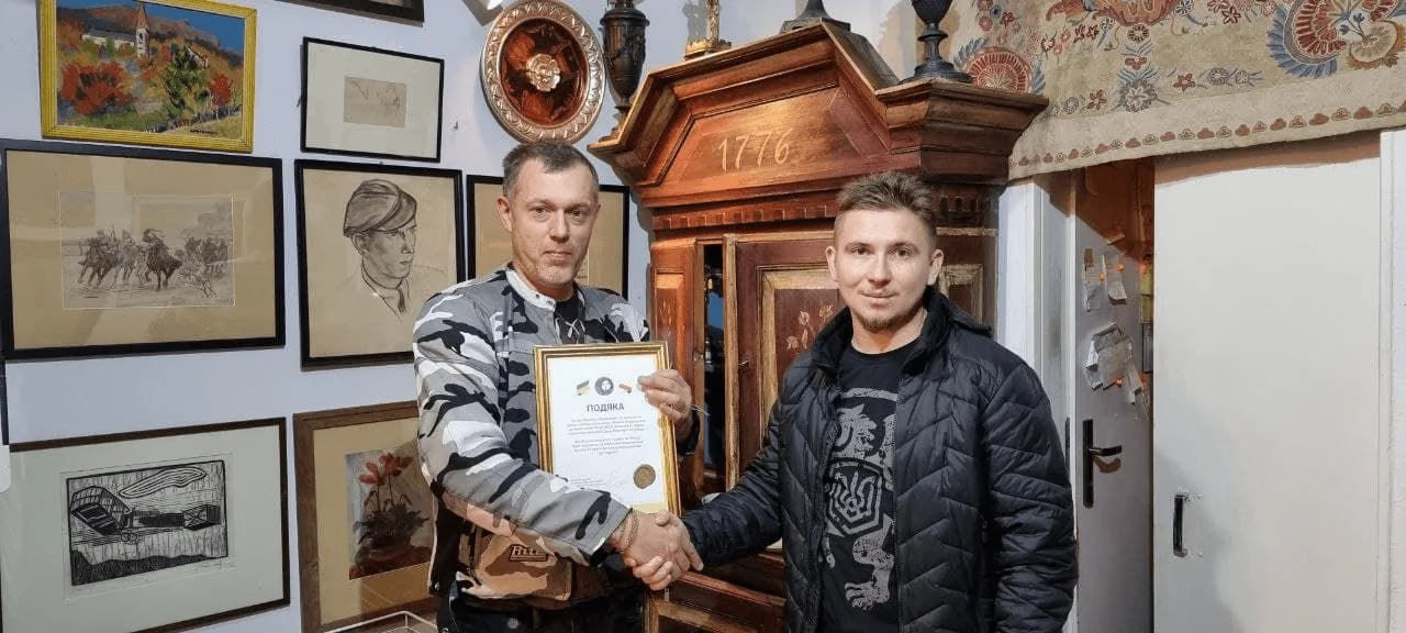 У кімнаті з картинами на стінах двоє чоловіків тиснуть один одному руки, один із них тримає грамоту. Позаду видно старовинні меблі та оформлену експозицію з малюнками.