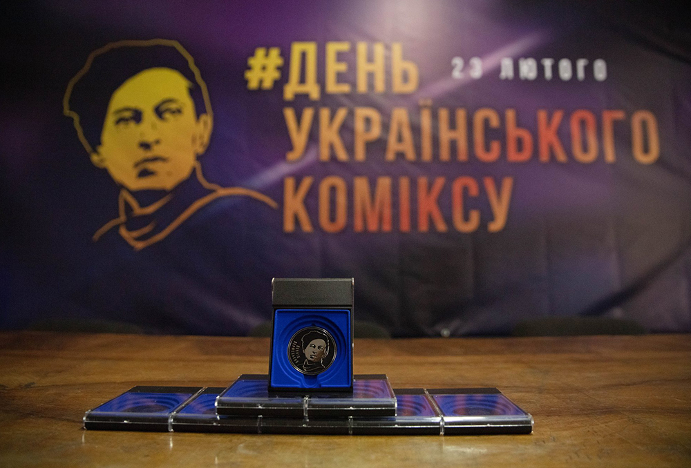 На столі розміщено пам’ятну монету у футлярі та кілька капсул, а на тлі видно банер із написом «День українського коміксу» і датою 23 лютого. Ліворуч на банері зображено стилізований портрет у жовтих тонах.