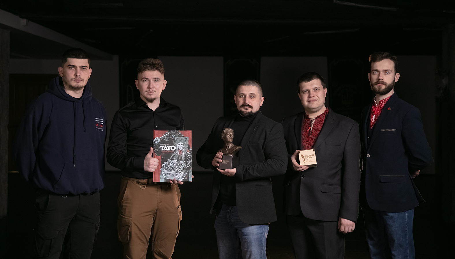 П’ятеро чоловіків стоять у темному приміщенні, дивлячись у камеру, дехто тримає книгу, статуетку та коробку з відзнакою. Вони позують разом, утворюючи груповий портрет, пов’язаний із коміксами або нагородженням.