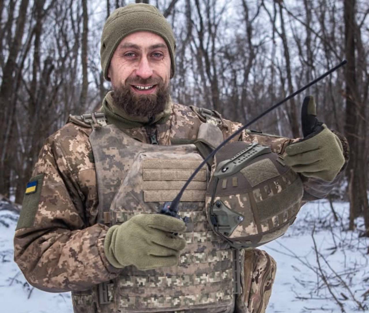 Боєць ЗСУ в бронежилеті, з рацією в руках посміхається.