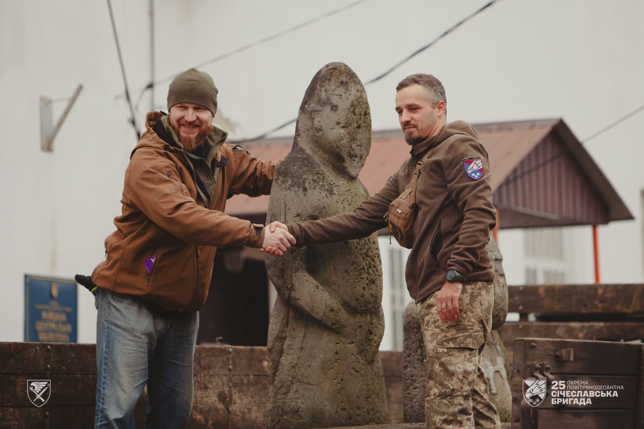 Двоє чоловіків тиснуть один одному руки, стоячи поруч зі стародавньою кам’яною статуєю просто неба. Один із них у військовій формі з шевроном підрозділу, інший у цивільному одязі, і обидва виглядають задоволеними під час цієї символічної зустрічі.