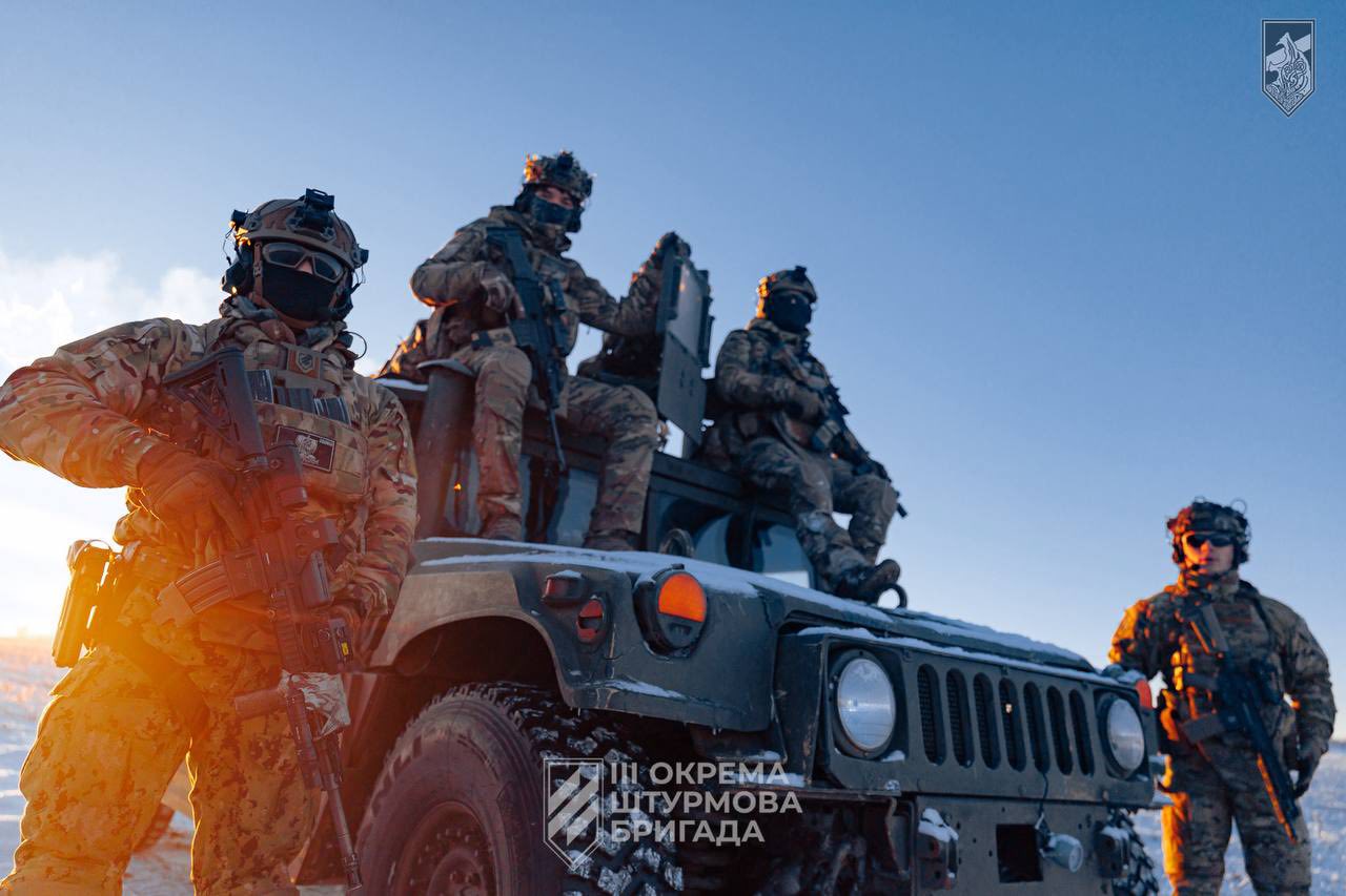 Четверо військових у повному спорядженні перебувають біля броньованого автомобіля на тлі зимового пейзажу під час заходу сонця. Троє з них розмістилися на машині, тримаючи зброю напоготові, ще один стоїть поруч, а тепле світло сонця підсвічує їхні силуети.