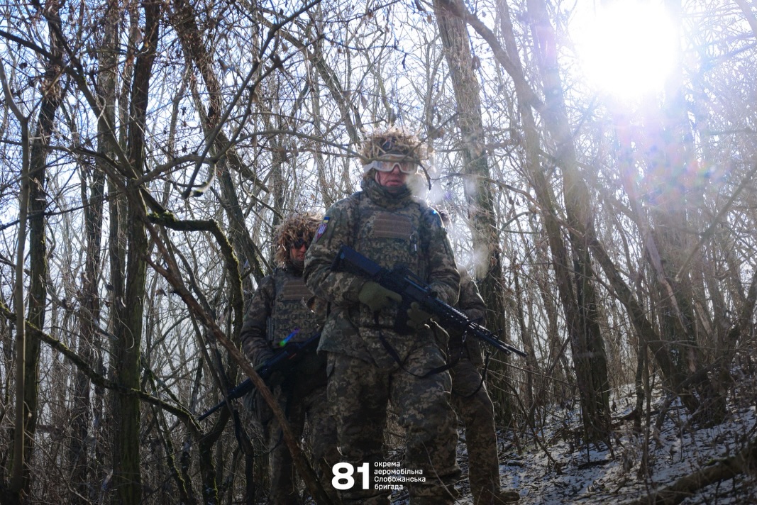 Група військовослужбовців у камуфляжі рухається крізь зимовий ліс, тримаючи напоготові автомати. Сонячне світло пробивається крізь голі гілки дерев, підсвічуючи їхнє спорядження та холодний засніжений ландшафт.