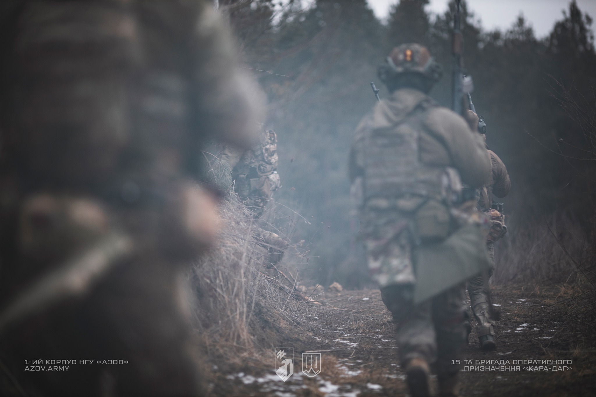 Озброєні військовослужбовці у спорядженні рухаються вузькою лісовою стежкою в холодну пору, тримаючи зброю напоготові. Кадр знятий з невеликої дистанції зі збереженням розмиття на передньому плані, що підкреслює рух групи крізь туманний ліс.