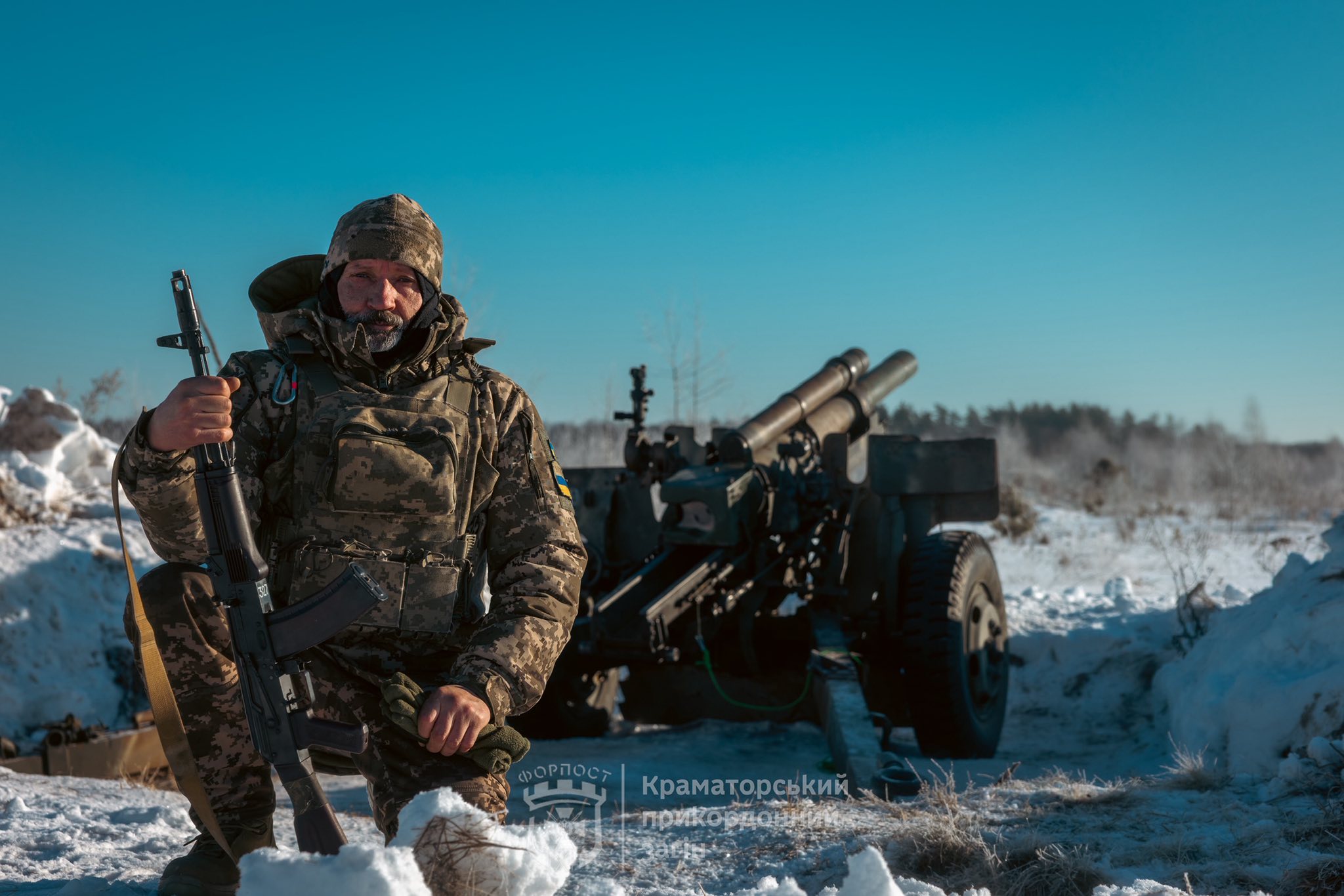 Військовий у зимовому камуфляжі стоїть на засніженій позиції, тримаючи автомат, а позаду нього розгорнута польова гармата. Морозний пейзаж і ясне небо підкреслюють фронтову обстановку та готовність підрозділу до роботи.
