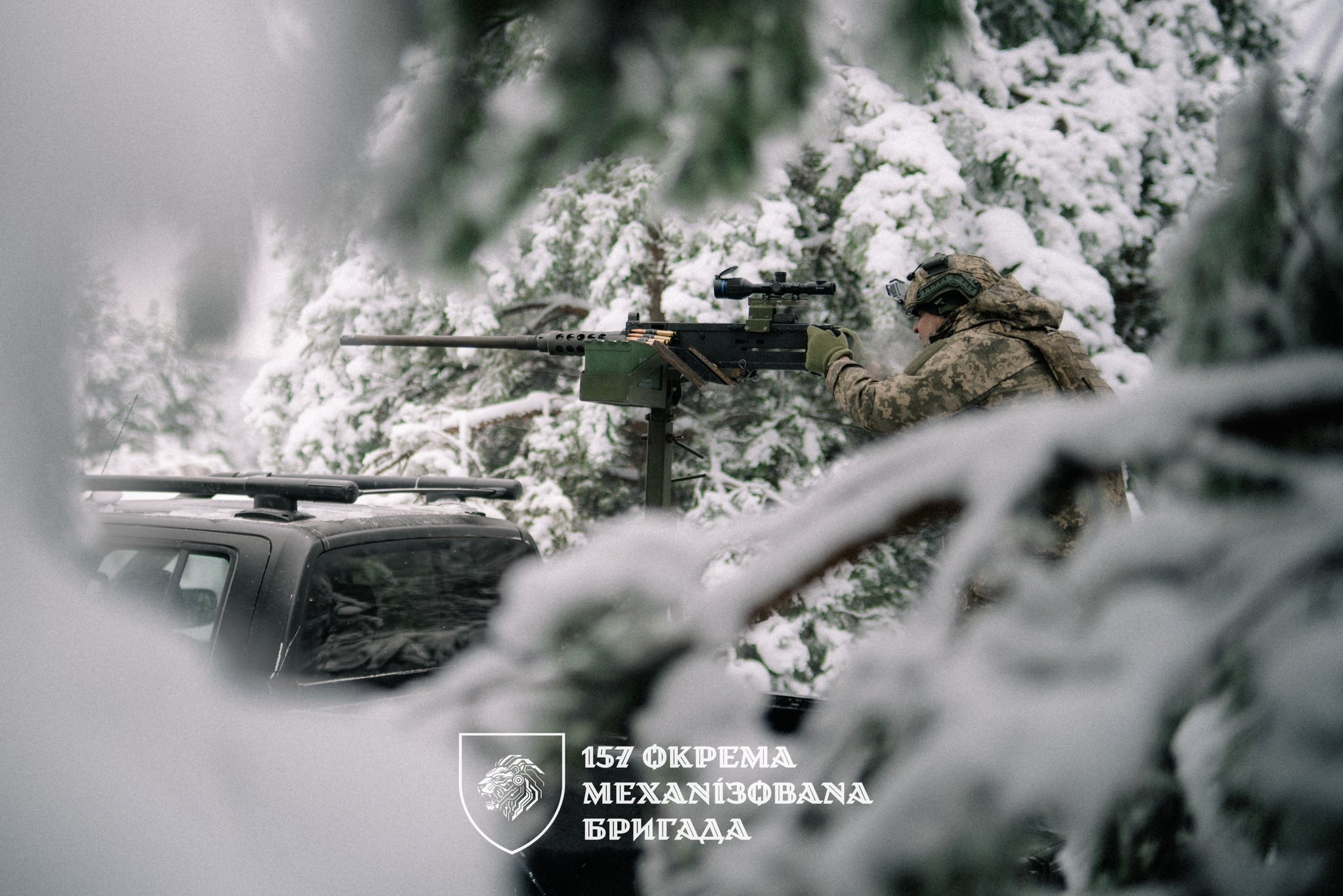 Військовослужбовець у зимовому лісі веде спостереження, працюючи з кулеметом, встановленим на пікапі, серед засніжених гілок і дерев. У нижній частині зображення розміщено емблему та напис 157-ї окремої механізованої бригади.