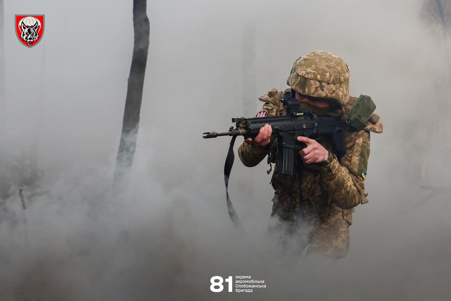 Військовослужбовець у камуфляжі веде прицільний вогонь з автомата, просуваючись крізь густий дим. Обличчя частково прикрите, а навколо панує напружена атмосфера бойових дій.
