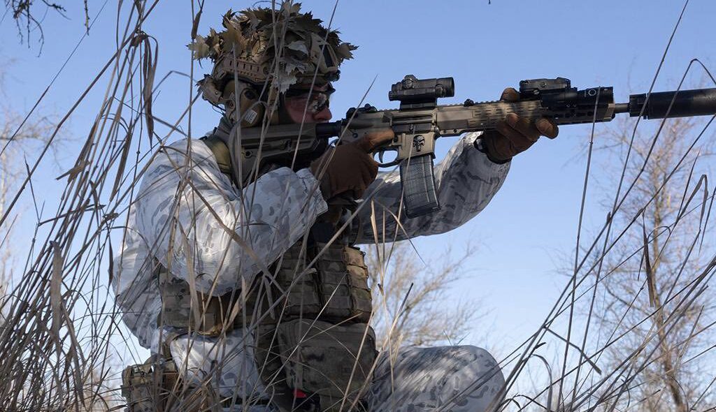Боєць у зимовому камуфляжі, сховавшись серед сухої трави та гілля, прицільно веде спостереження зі штурмовою гвинтівкою, стоячи на одному коліні. На шоломі видно маскувальні елементи, а внизу зображення розміщено емблему підрозділу «Омега» Центру спеціального призначення.