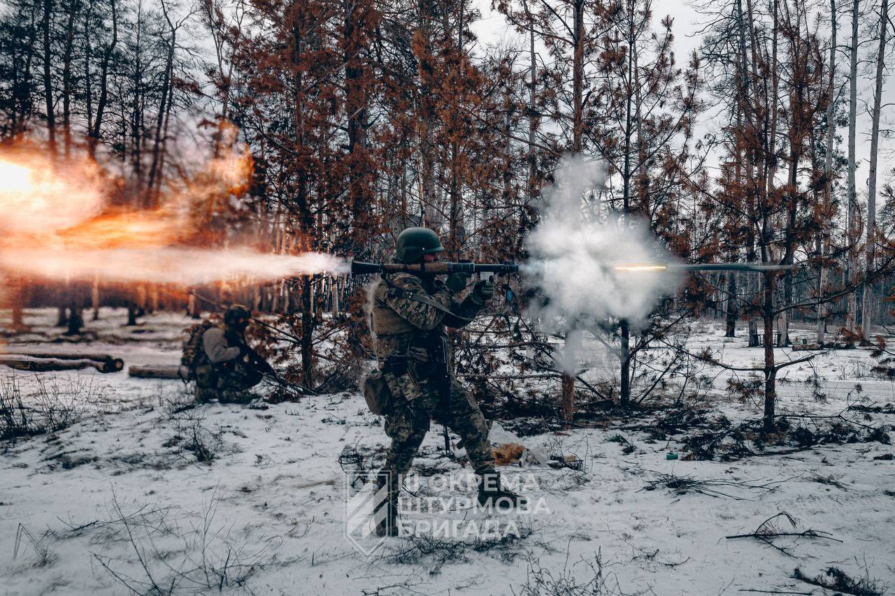 Військовий у зимовому лісі веде вогонь із ручного протитанкового гранатомета, з якого виривається яскравий спалах і дим. Позаду нього інший боєць перебуває напоготові серед засніжених дерев і обгорілої рослинності.