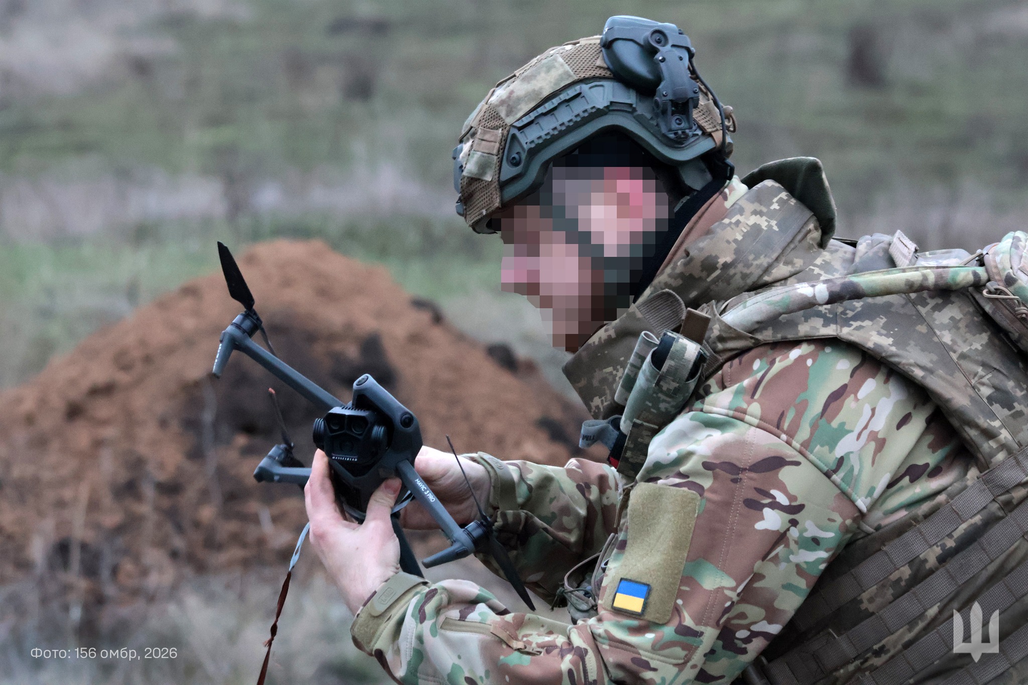 Військовослужбовець у камуфляжі та шоломі тримає в руках невеликий безпілотник, готуючи його до запуску або перевіряючи перед польотом. На рукаві видно український прапор, а позаду — польова місцевість із насипом землі, що нагадує позицію або укриття.
