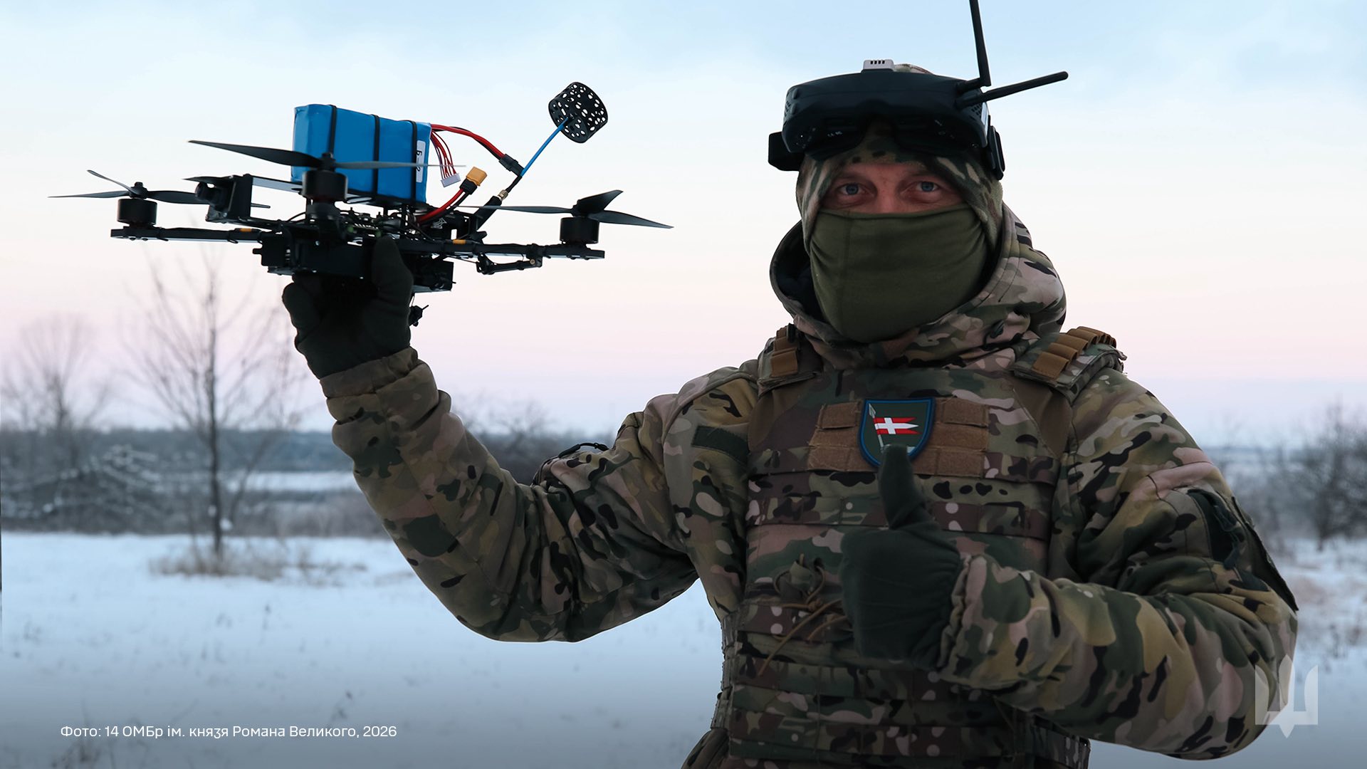 Військовослужбовець у зимовій формі та захисних окулярах стоїть на засніженій місцевості, тримаючи в руці квадрокоптер із встановленим обладнанням. Іншою рукою він показує жест «лайк», а на грудях видно шеврон підрозділу.