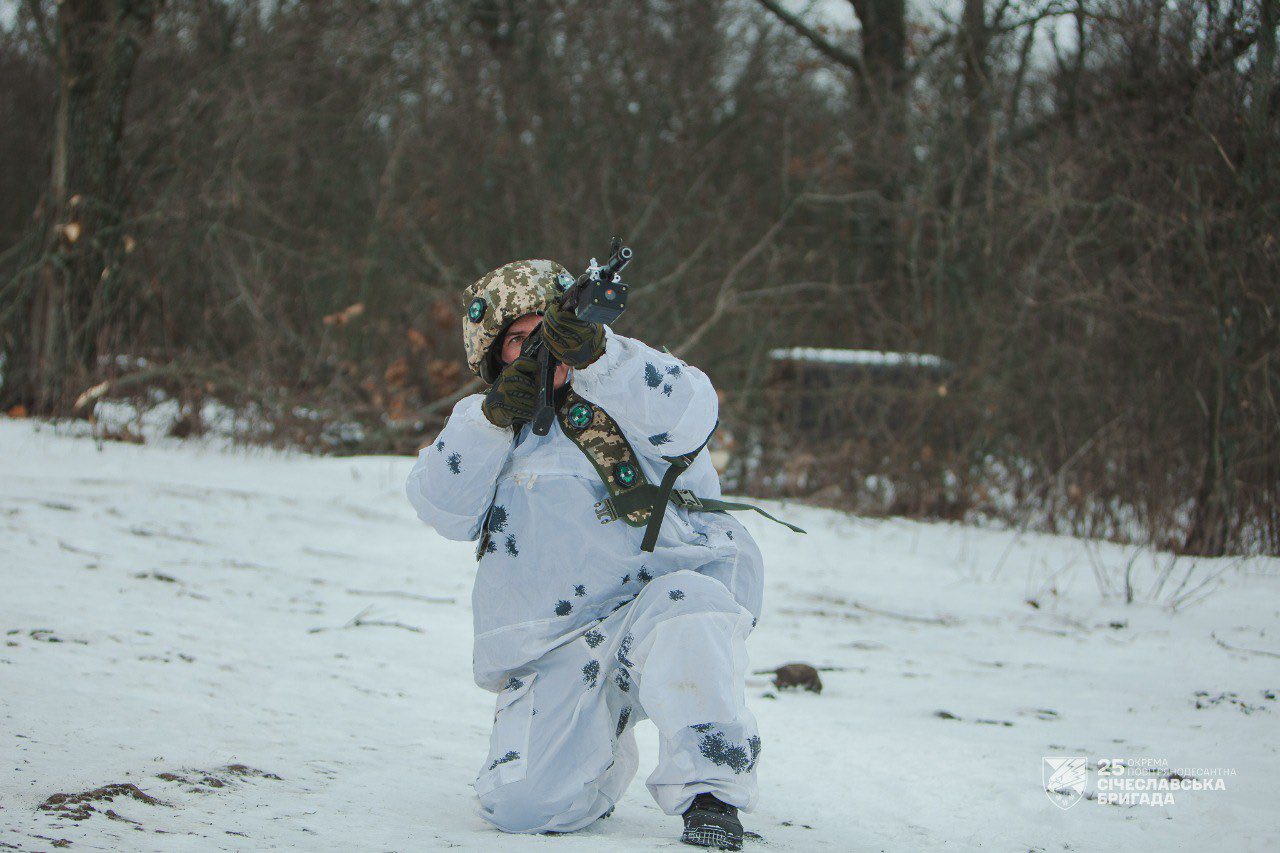 Військовослужбовець у зимовому маскувальному костюмі стоїть на коліні посеред засніженого лісу та веде прицільний вогонь з автомата. На його шоломі й спорядженні видно шеврони, а на тлі — оголені дерева й холодний зимовий пейзаж.