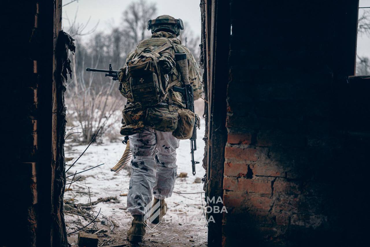 Військовий із великим наплічником і кулеметною стрічкою виходить через пролом у зруйнованій будівлі назовні. Попереду видно засніжену місцевість і голі дерева, що підкреслює зимові умови та фронтову обстановку.