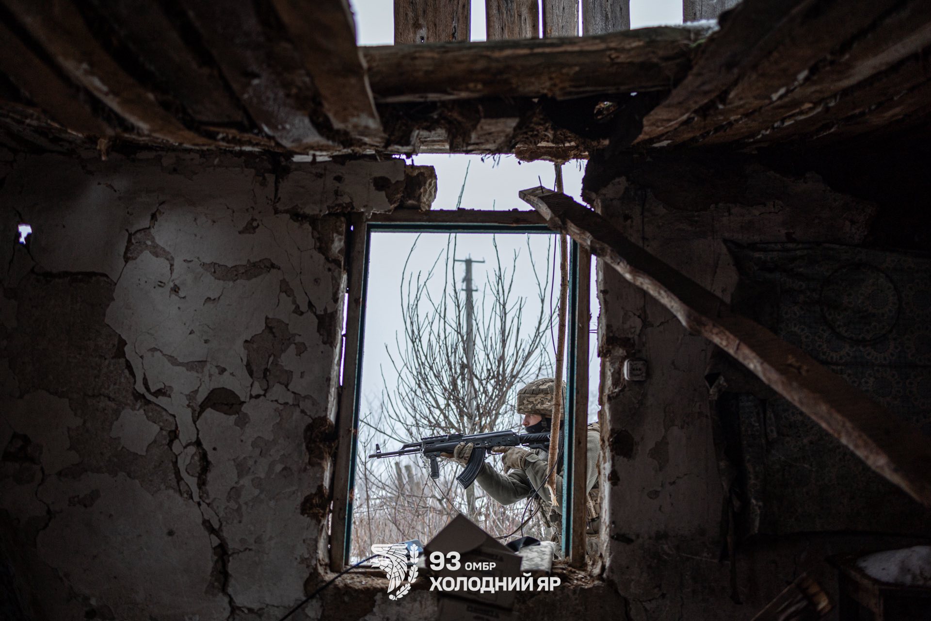 Через віконний проріз зруйнованої будівлі видно військового зі зброєю, який веде спостереження або прикриття на вулиці. Усередині — обвалена стеля й потріскані стіни, що підкреслюють сліди бойових руйнувань.