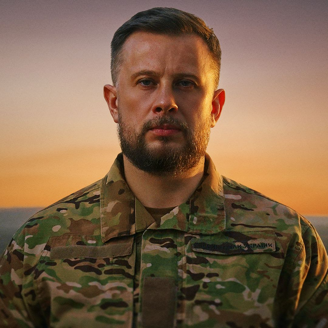 Чоловік у військовій формі дивиться просто в камеру на тлі теплого заходу сонця. Камуфляжний одяг і серйозний вираз обличчя створюють стриманий портретний образ.