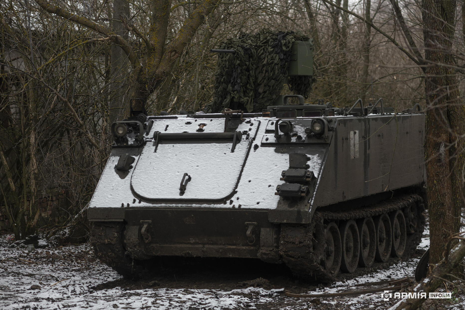 Гусенична броньована машина М113, замаскована сіткою, стоїть у лісовій місцевості серед голих дерев. На її передній броні видно тонкий шар снігу, а гусениці забруднені болотом, що свідчить про рух бездоріжжям. Атмосфера кадру передає холодну пору року й напружену, польову обстановку.