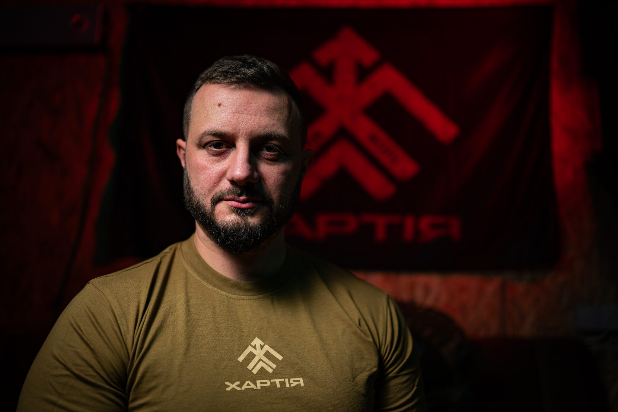 Чоловік у футболці з написом «Хартія» сидить у темному приміщенні, освітлений м’яким світлом. Позаду видно прапор із символом під червоним підсвічуванням.